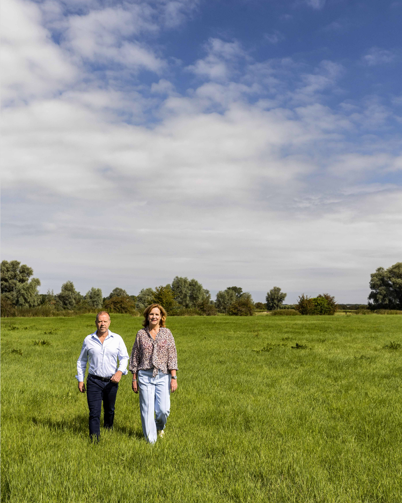 Peter van Veelen en Martine Goedegebuure lopen door een groen weiland onder een blauwe lucht