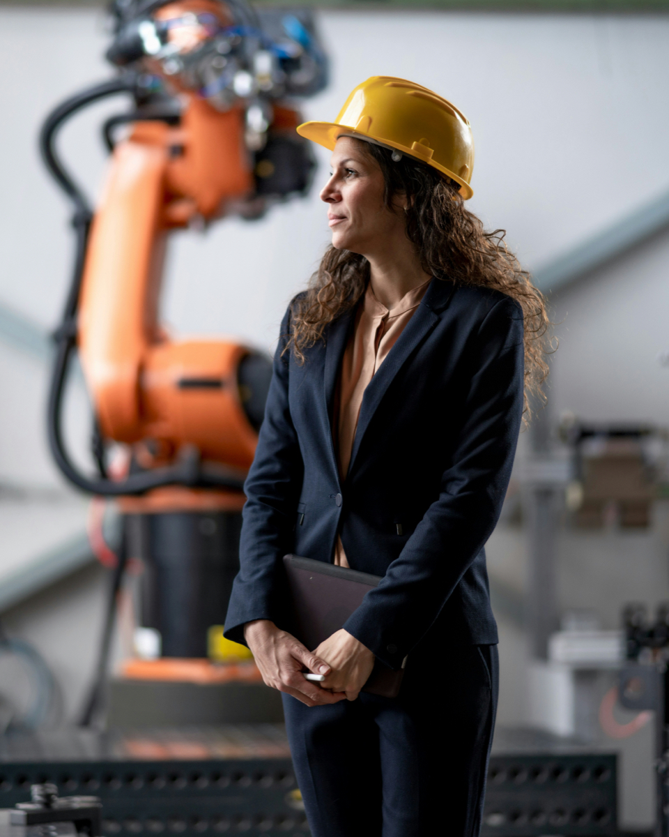 Zakenvrouw met gele veiligheidshelm in fabriekshal voor industriële robotarm, overweegt warmteterugwinning bij corrosieve rookgassen
