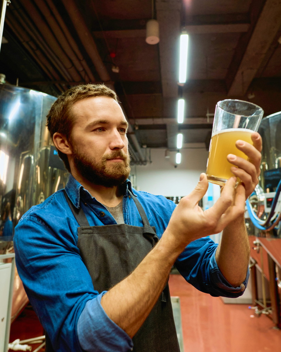 Brouwer inspecteert een glas met troebel bier in een moderne productieruimte, met glimmende tanks op de achtergrond.