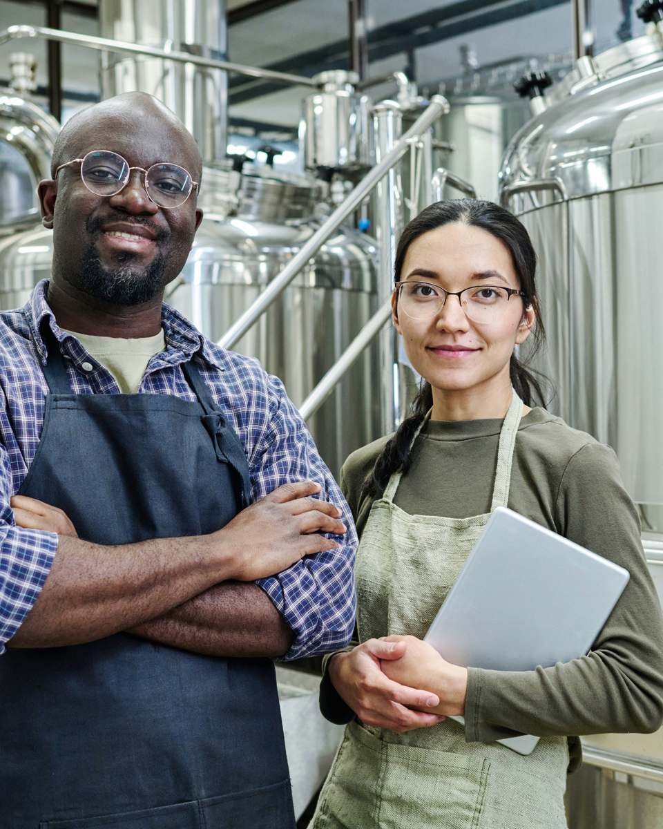 Twee medewerkers in een industriële installatie met roestvrijstalen tanks, dragen schorten en bekijken samen de uitkomsten van de Warmteterugwinning Scan.