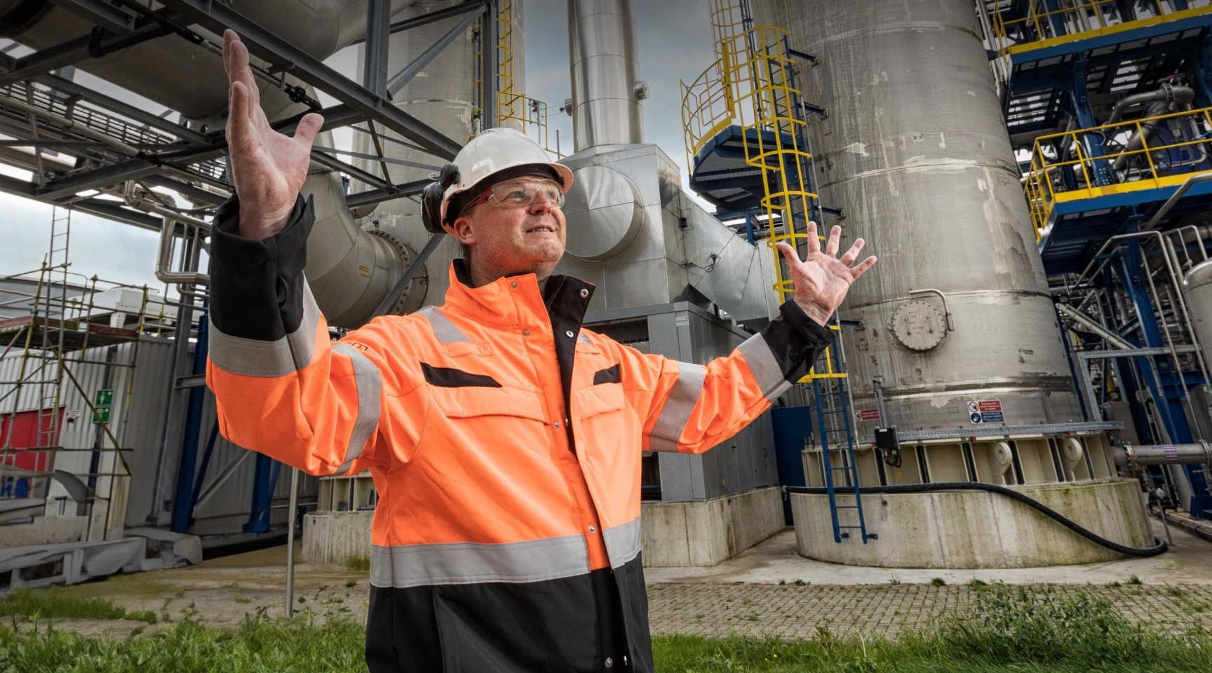 Engineer in oranje werkjas en helm staat trots voor een industriële installatie, symbool voor sterke samenwerkingen met partnerbedrijven.