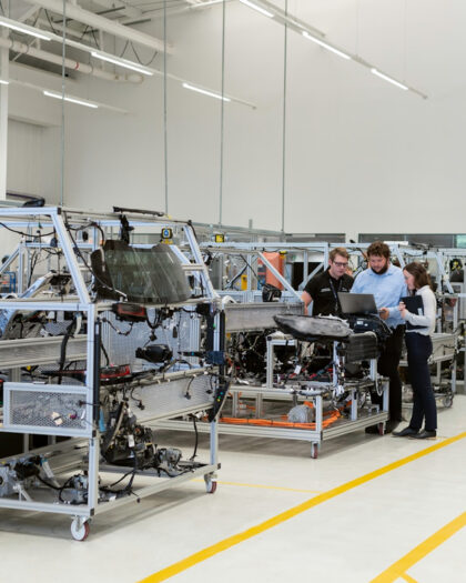 Drie ingenieurs overleggen bij een industriële testopstelling in een moderne productiehal, symbool voor technologische innovatie en samenwerking.