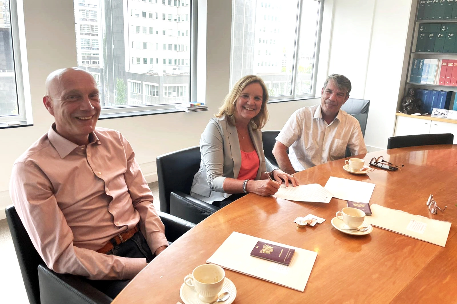 Martine Goedegebuure ondertekent samen met twee mannen de overname van Heat Matrix door Bos Nieuwerkerk aan een vergadertafel met uitzicht op de stad.