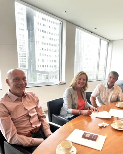 Martine Goedegebuure ondertekent samen met twee mannen de overname van Heat Matrix door Bos Nieuwerkerk aan een vergadertafel met uitzicht op de stad.