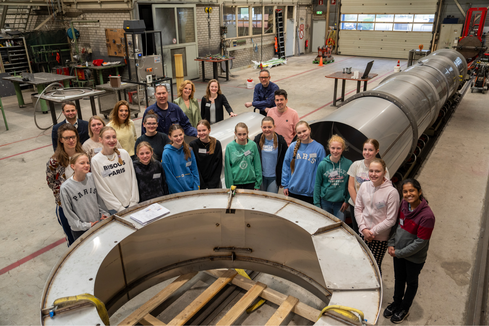 Groepsfoto van leerlingen en professionals in een industriële werkplaats naast een groot onderdeel van een warmtewisselaar.