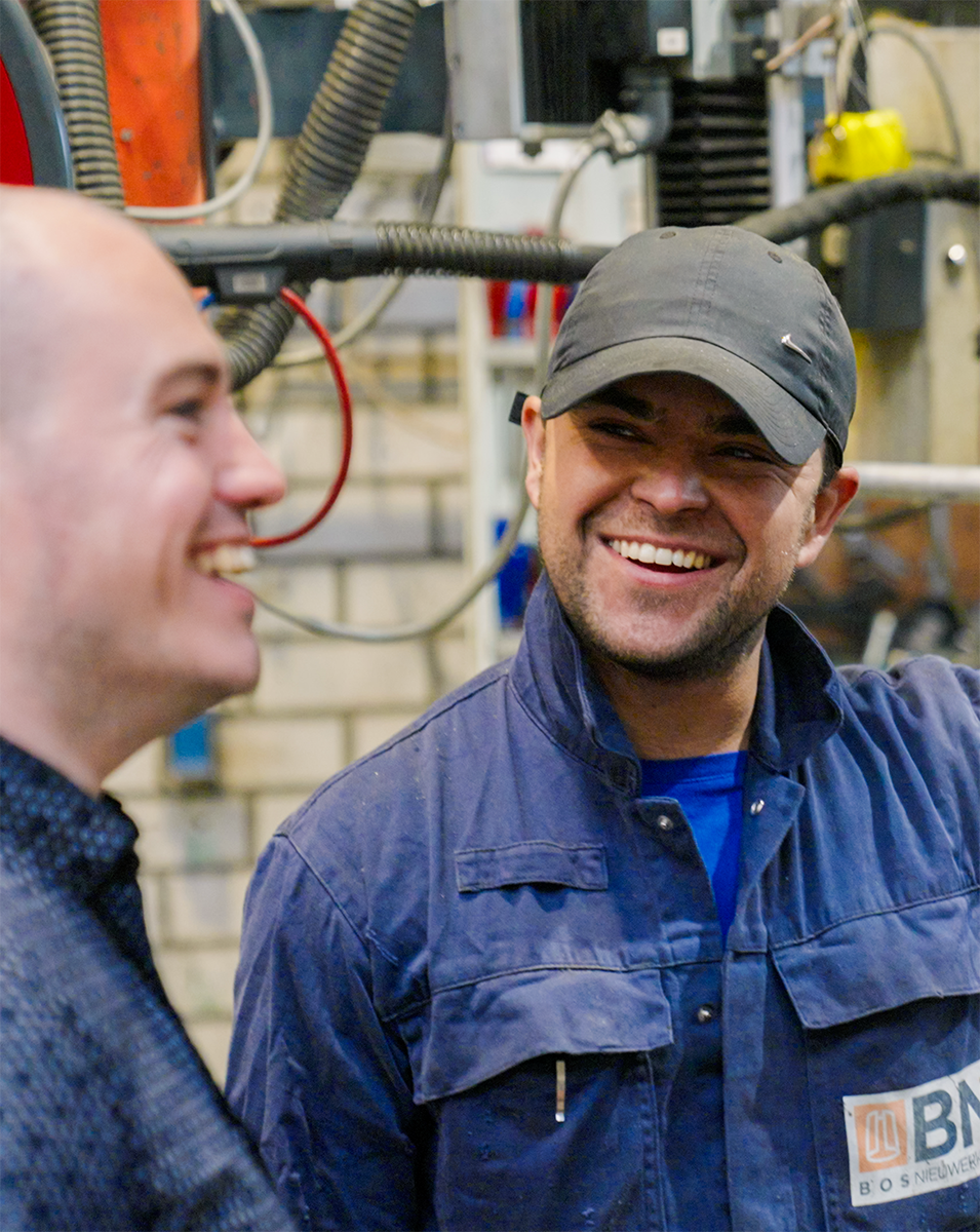 Smiling technician in a navy-blue work overall with a BN (Bos Nieuwerkerk) logo, standing beside a colleague in an industrial workshop.